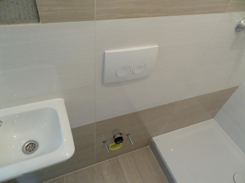 Wykończona łazienka: biała umywalka, podwieszana toaleta z przyciskiem i beżowe płytki. Widoczne elementy instalacji pod toaletą.