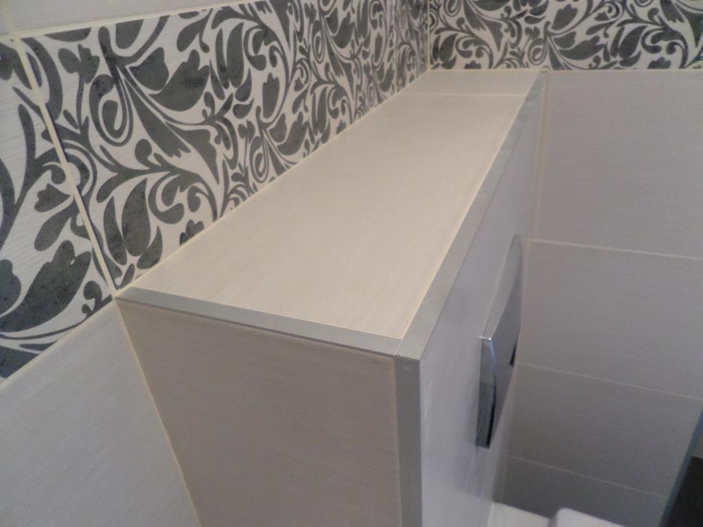 Wykończona łazienka: białe płytki, dekoracyjny wzór na ścianie, zabudowa toalety z aluminiowymi listwami wykończeniowymi i przyciskiem spłukiwania.