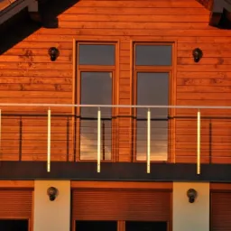 Fragment drewnianego domu z balkonem zabezpieczonym nowoczesną balustradą z cienkich, metalowych linek i pionowych, jasnych słupków, oświetlonego ciepłym, zachodzącym słońcem.