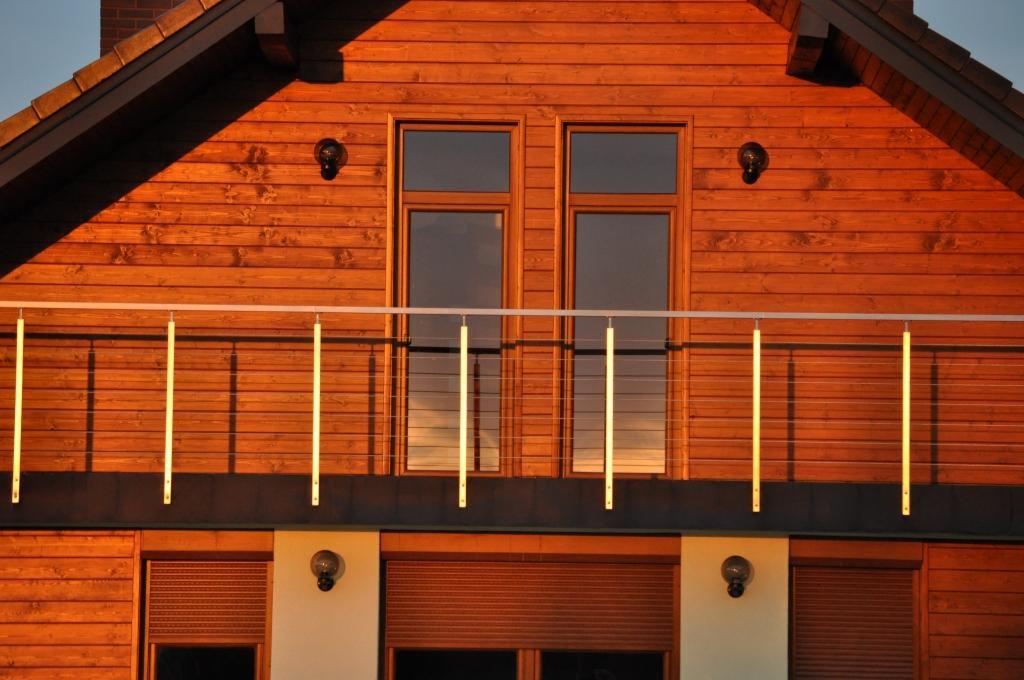 Fragment drewnianego domu z balkonem zabezpieczonym nowoczesną balustradą z cienkich, metalowych linek i pionowych, jasnych słupków, oświetlonego ciepłym, zachodzącym słońcem.