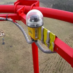 Czerwona konstrukcja z zamontowanym żółtym oświetleniem przeszkodowym z przezroczystą kopułą, widok z góry na krajobraz z pozostałościami budynku.