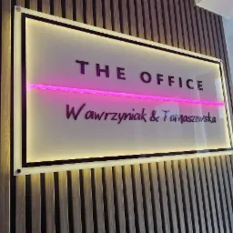Podświetlany szyld firmy 'The Office Wawrzyniak & Tomaszewska' na drewnianej ścianie, oświetlony różową neonową linią.