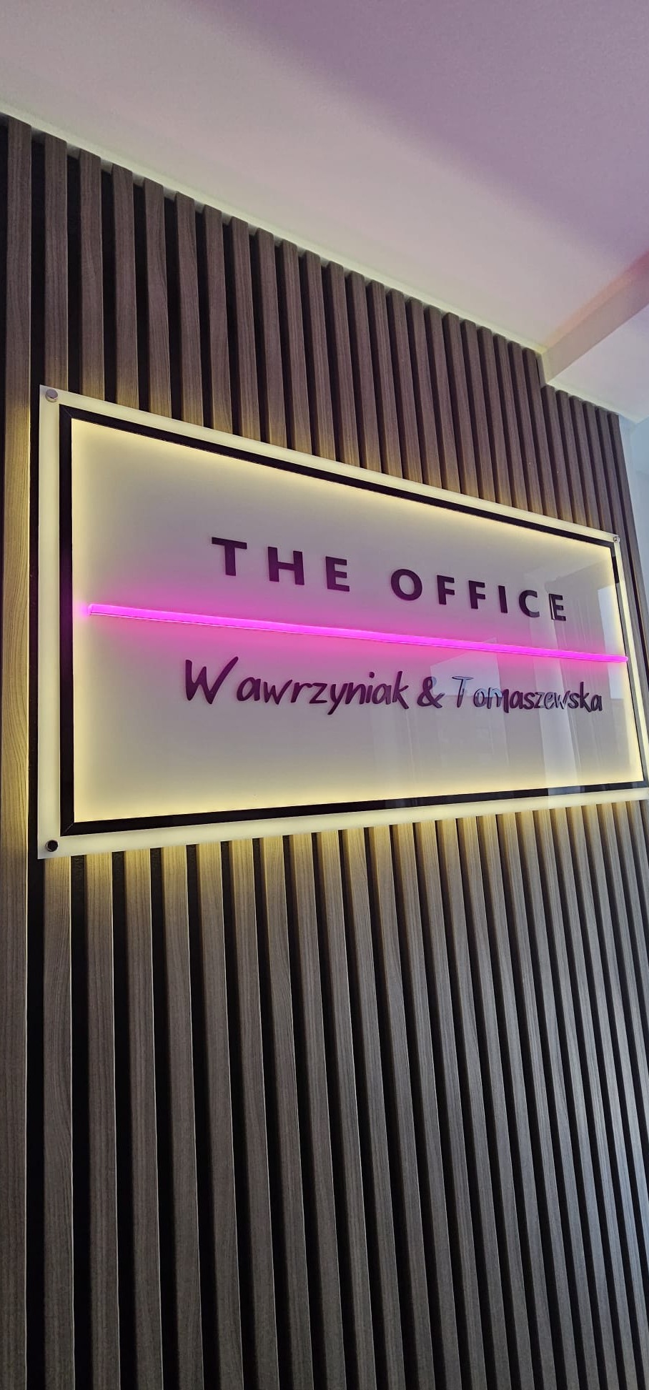 Podświetlany szyld firmy 'The Office Wawrzyniak & Tomaszewska' na drewnianej ścianie, oświetlony różową neonową linią.