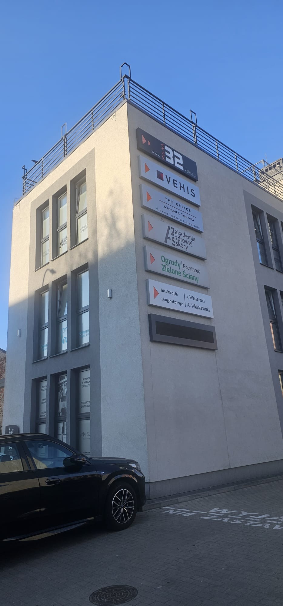 Biurowiec z tablicami informacyjnymi firm, w tym 'THE OFFICE' oferującej finanse, ubezpieczenia i biznes. Na parterze widoczne okna z reklamami. Częściowo widoczny czarny samochód.