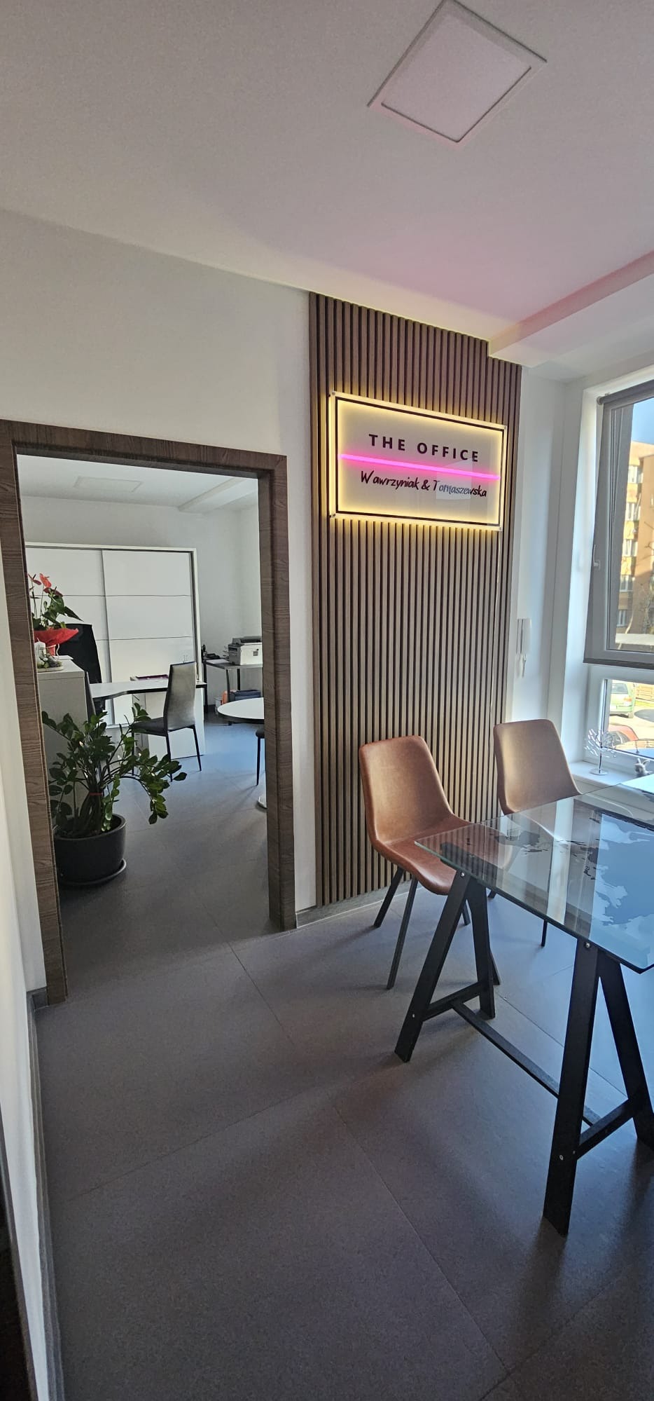 Nowoczesne biuro z drewnianą ścianą i podświetlanym logo 'The Office Wawrzyniak & Tomaszowska'. W tle widoczne biurka i krzesła, a także roślina doniczkowa.