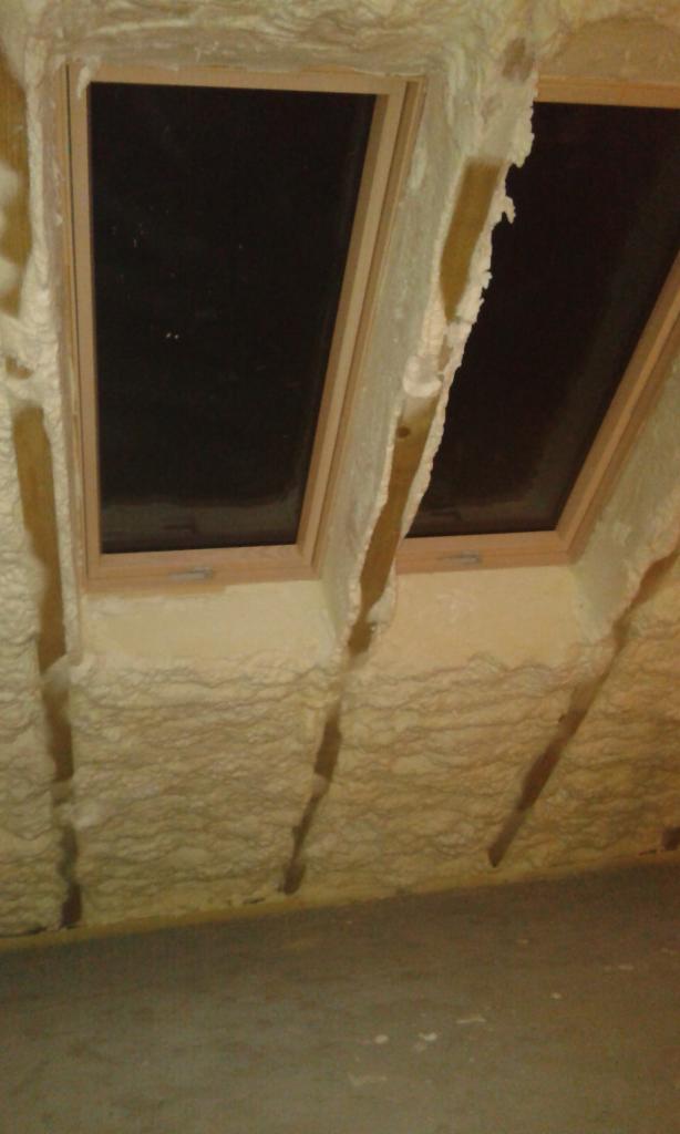 Dwa okna dachowe z drewnianymi ramami, obficie otoczone pianką izolacyjną PUR, wypełniającą przestrzenie wokół nich. Pianka pokrywa również fragmenty ściany i sufitu.