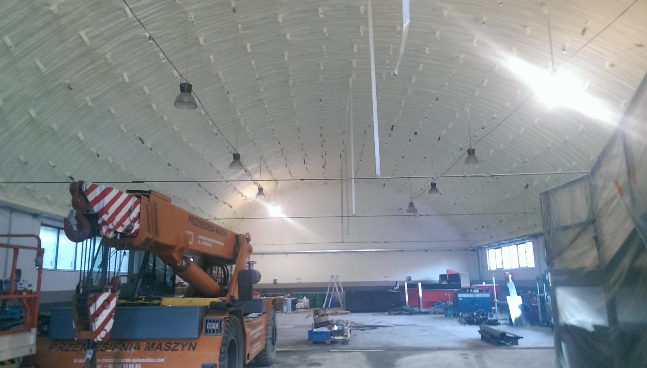 Wnętrze hali z izolacją pianką PUR na sklepieniu, widoczny dźwig. Biała pianka tworzy wzór na suficie. Lampy wiszące, okna w tle. Praca w toku.