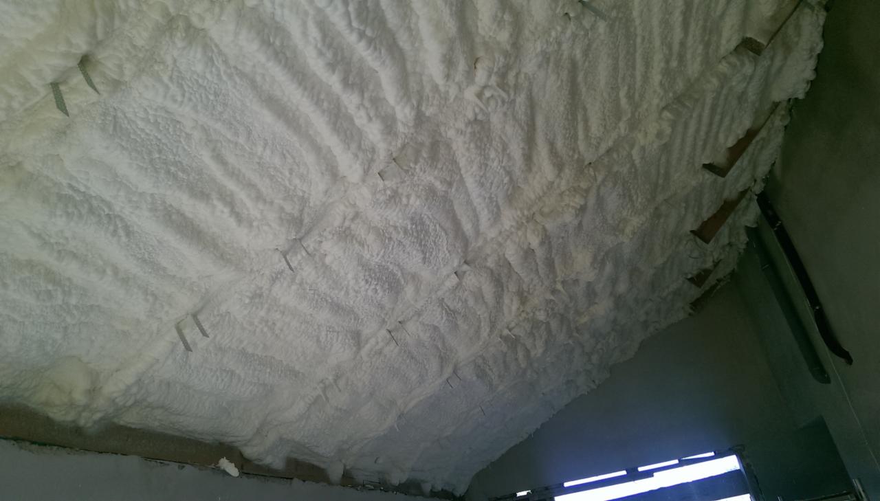 Biała pianka PUR zaaplikowana na poddasze, widoczne metalowe elementy mocujące. Izolacja natryskowa, nierówna powierzchnia piany.