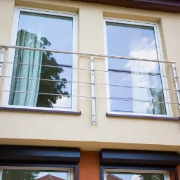 Balkon z beżową elewacją i nowoczesną, minimalistyczną balustradą ze stali nierdzewnej, zamontowaną na tle dwóch okien z roletami zewnętrznymi.