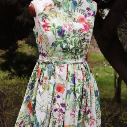 Letnia sukienka z motywem florystycznym na manekinie, z dekoracyjną ważką na ramieniu, na tle zieleni. Wzór w kwiaty i liście w odcieniach zieleni, różu i fioletu.