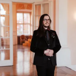 Elegancki mężczyzna w czarnym, aksamitnym garniturze, z butonierką z ciemnej róży, pozuje w jasnym wnętrzu z otwartymi, przeszklonymi drzwiami.