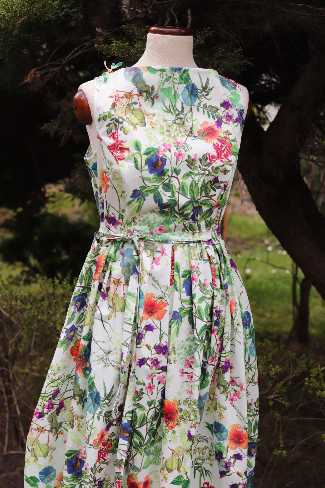 Letnia sukienka z motywem florystycznym na manekinie, z dekoracyjną ważką na ramieniu, na tle zieleni. Wzór w kwiaty i liście w odcieniach zieleni, różu i fioletu.