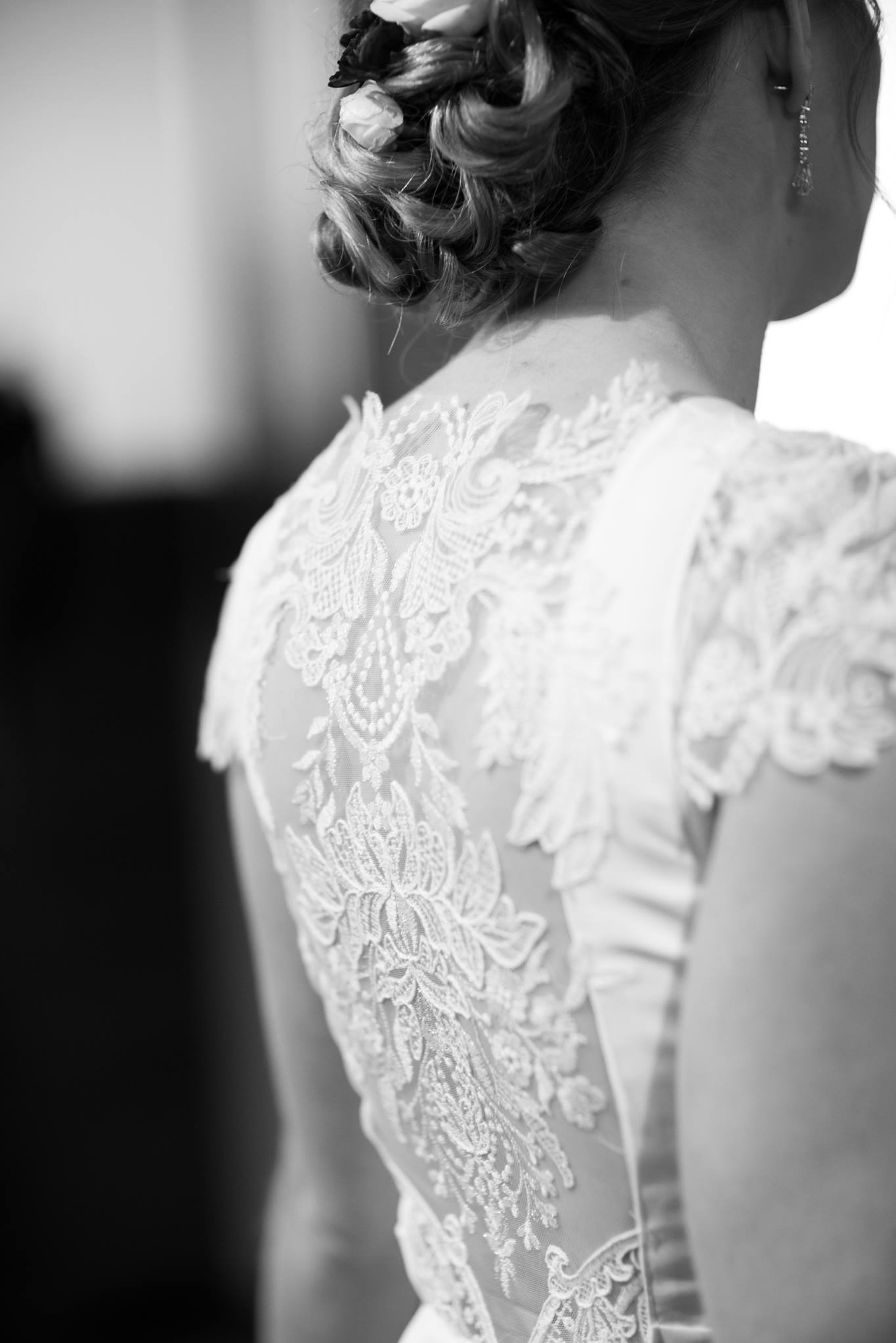 Czarno-białe zbliżenie na plecy kobiety w koronkowej sukni, z delikatnymi kwiatami we włosach i subtelnymi kolczykami. Widoczny misterny wzór koronki.