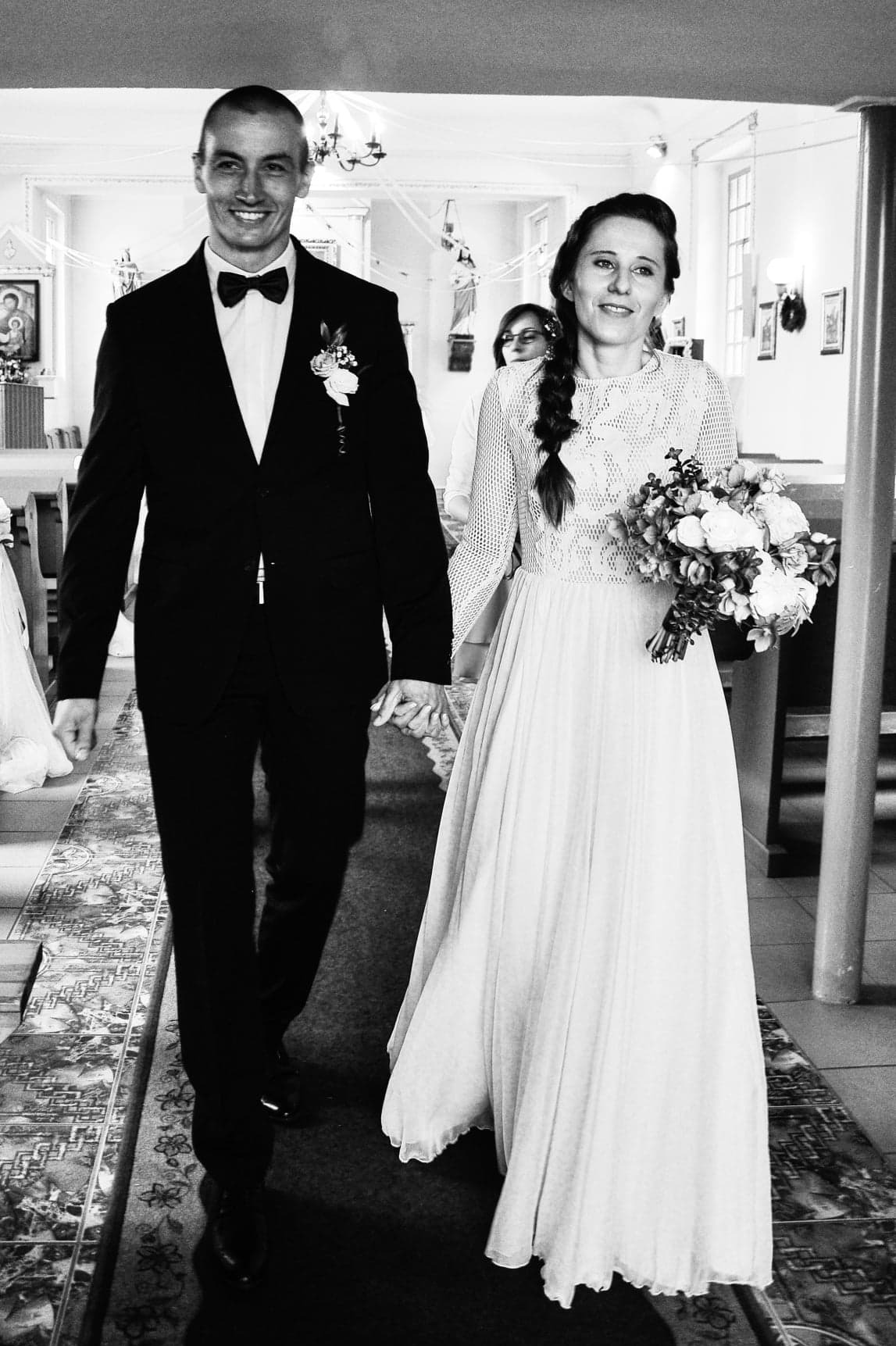 Para młoda w kościele: panna młoda w koronkowej sukni z bukietem, pan młody w smokingu. Czarno-białe zdjęcie ślubne z detalami wystroju wnętrza kościoła.