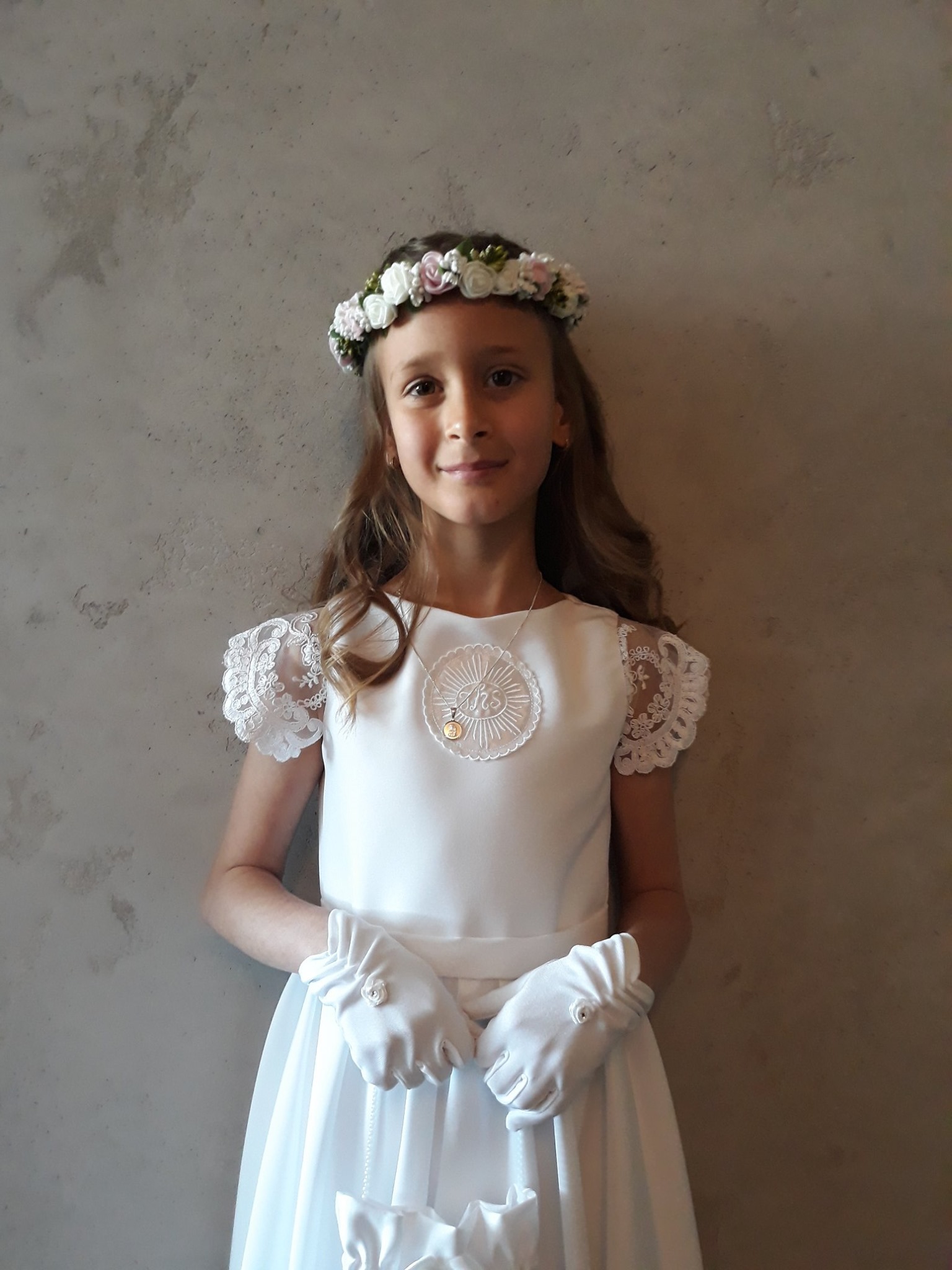 Dziewczynka w białej sukience komunijnej z wiankiem i rękawiczkami, stojąca na tle szarej ściany. Delikatne zdobienia z koronki i kwiatów, subtelna biżuteria.