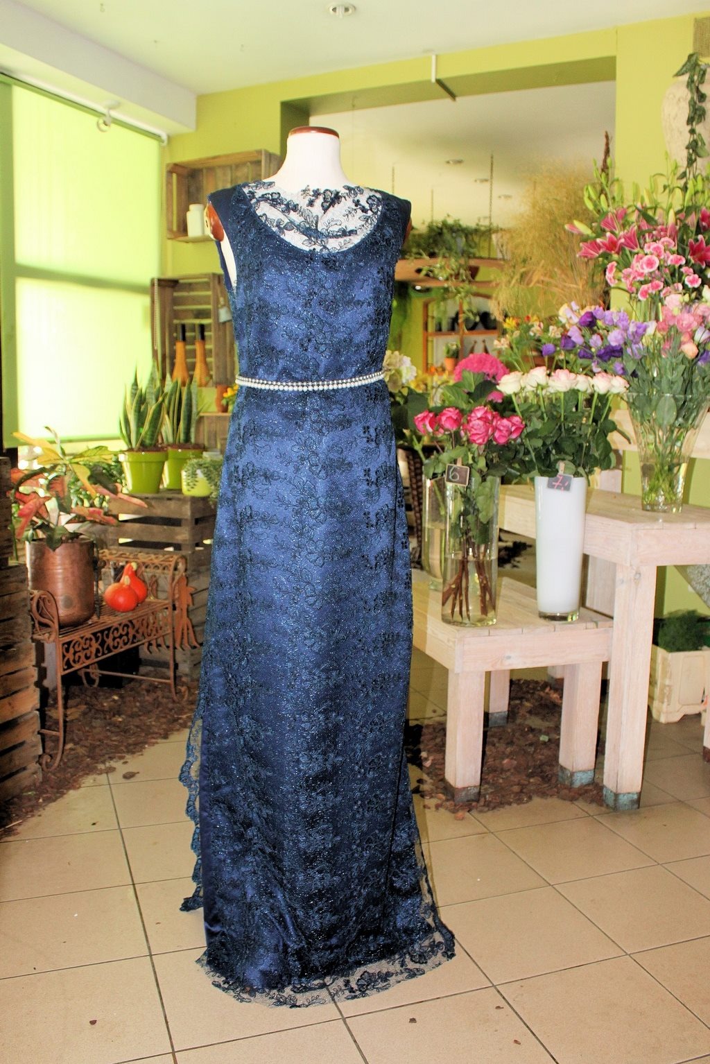 Elegancka, długa suknia wieczorowa w odcieniach granatu z koronkowymi zdobieniami, prezentowana na manekinie w otoczeniu kwiatów.