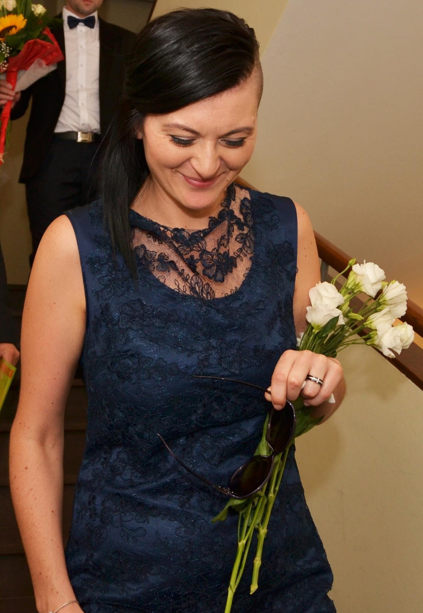Uśmiechnięta kobieta w granatowej koronkowej sukience, trzymająca okulary i bukiet białych kwiatów, z mężczyzną w tle.