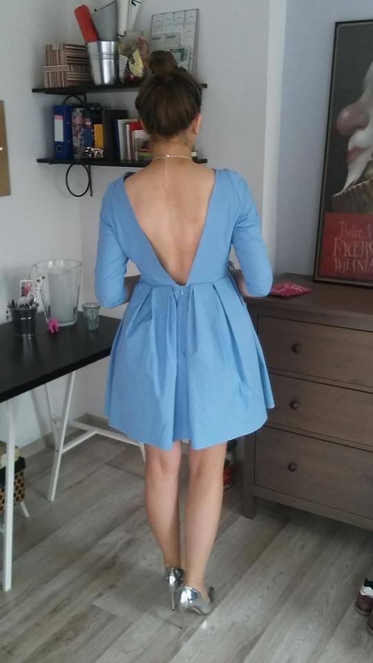 Jasnoniebieska, rozkloszowana sukienka z odkrytymi plecami, widok z tyłu, srebrne buty na obcasie, drewniana podłoga, komoda i półki w tle.