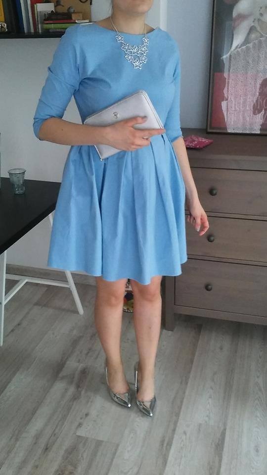 Kobieta w błękitnej sukience z plisowanym dołem, srebrnych butach i z srebrną torebką, prezentująca stylizację na tle komody i fragmentu stołu.