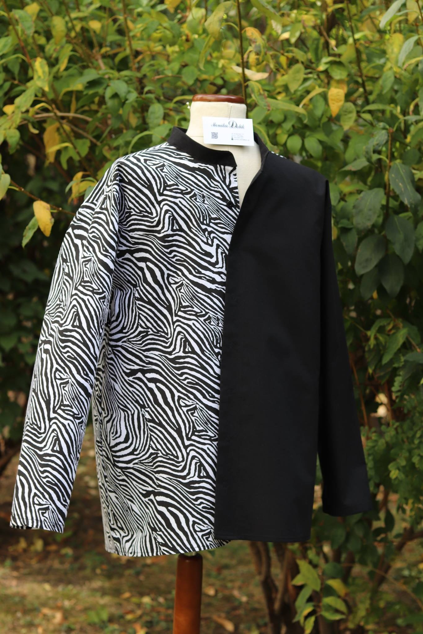 Dwukolorowa, asymetryczna koszula z długim rękawem, wzór zebra po lewej, gładka czerń po prawej, prezentowana na manekinie na tle zieleni.