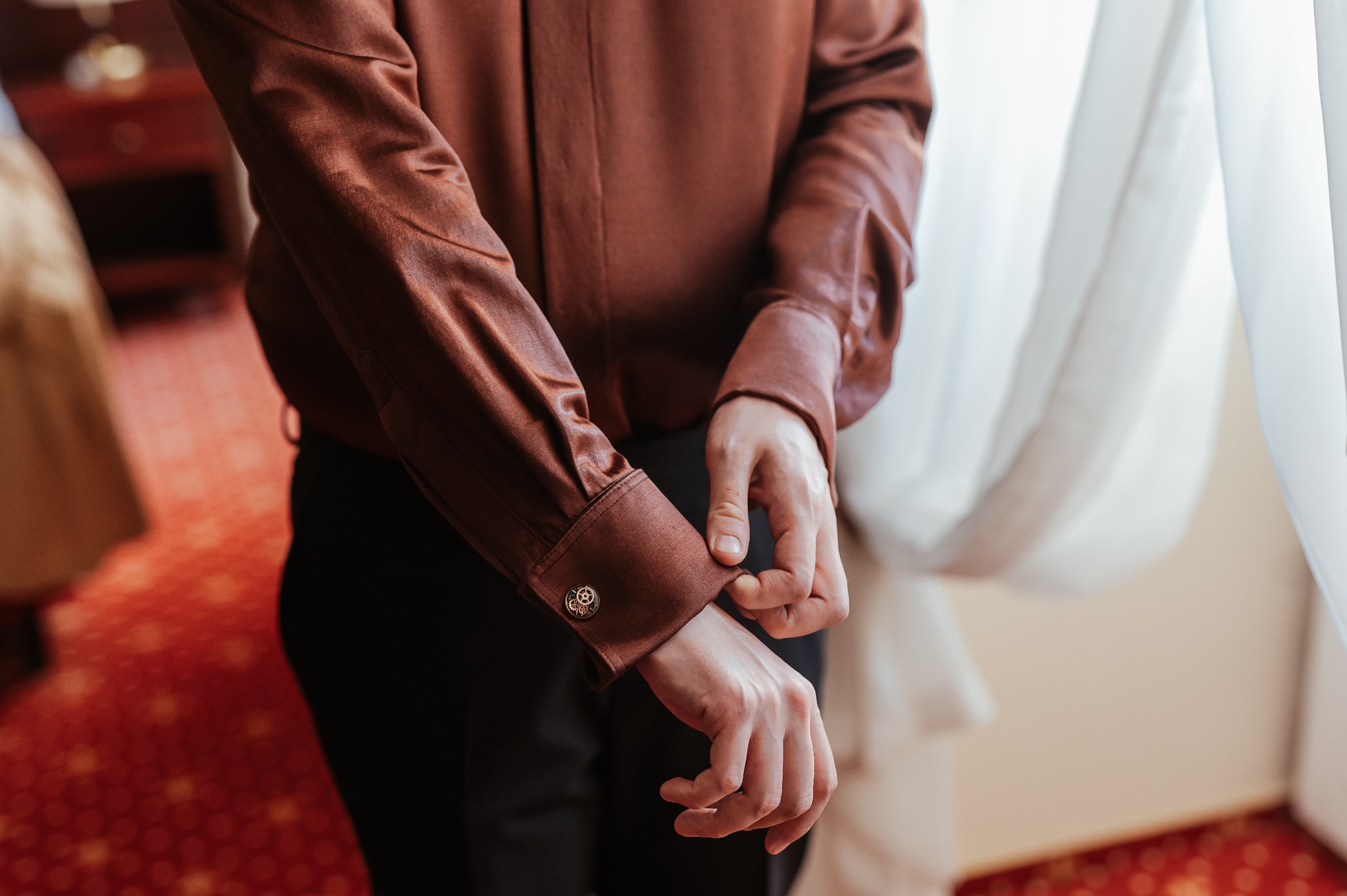 Brązowa, połyskująca koszula z mankietami zapinanymi na ozdobne spinki. Widoczne dłonie zapinające mankiet. Elegancki detal odzieży męskiej na tle okna.