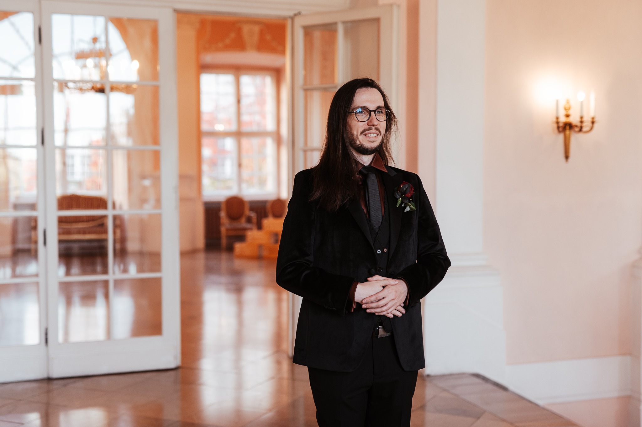 Elegancki mężczyzna w czarnym, aksamitnym garniturze, z butonierką z ciemnej róży, pozuje w jasnym wnętrzu z otwartymi, przeszklonymi drzwiami.