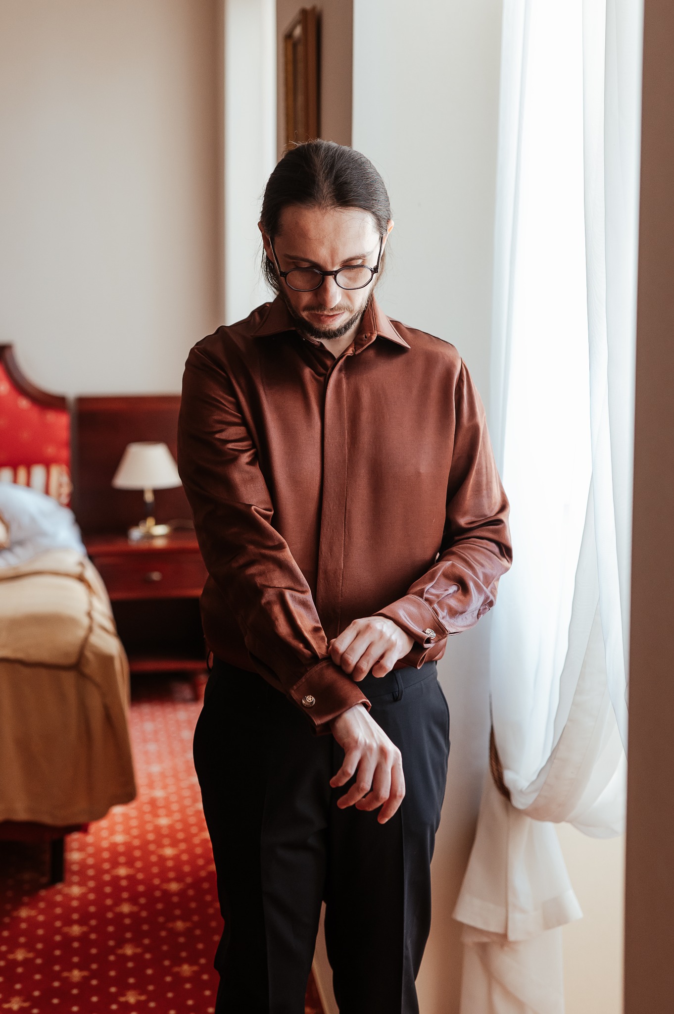 Mężczyzna w brązowej, satynowej koszuli zapina mankiet na tle okna z białymi zasłonami i czerwonej sypialni. Elegancki styl, dopasowany strój.