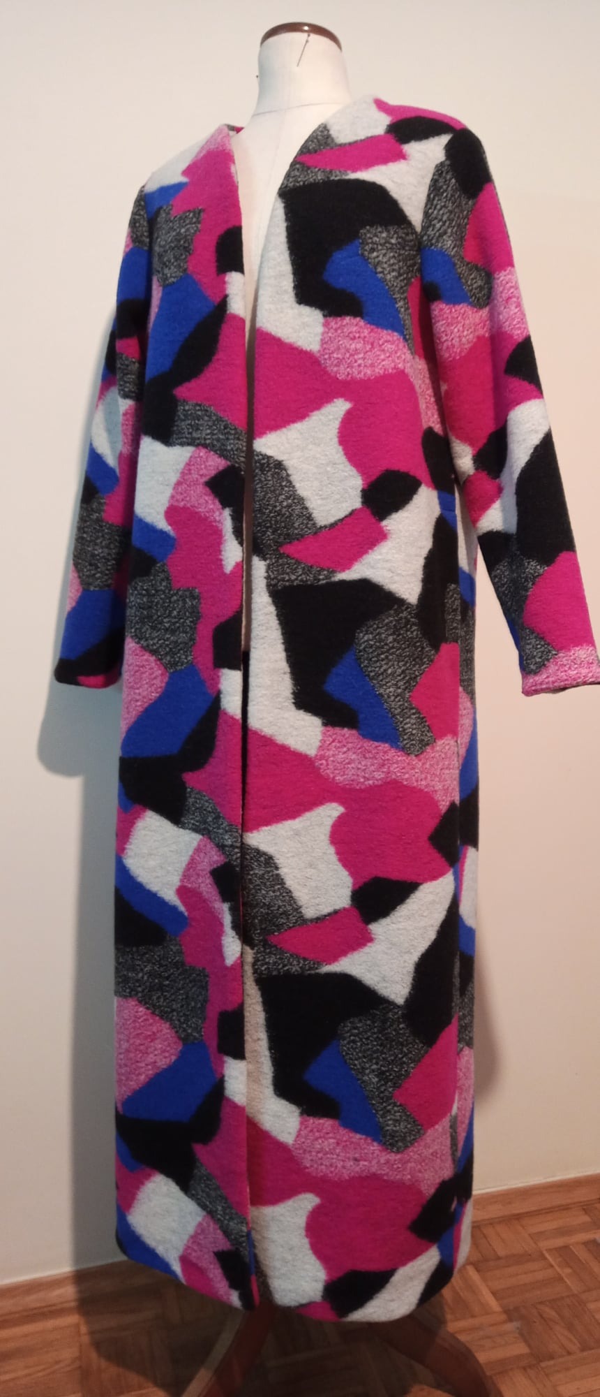 Długi, kolorowy płaszcz z geometrycznym wzorem, w odcieniach różu, błękitu, czerni i szarości, zaprezentowany na manekinie krawieckim.