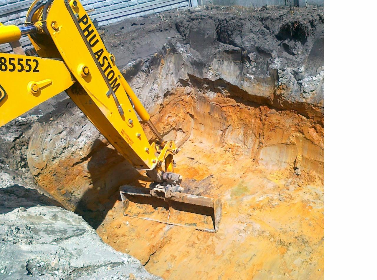 Żółta koparka z widocznym numerem seryjnym 85552 zagłębia się łyżką w ziemię, tworząc wykop z warstwami gleby o różnej kolorystyce: ciemnej, szarej i pomarańczowej.