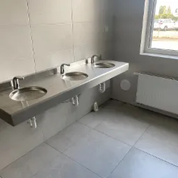 Wnętrze łazienki publicznej z trzema umywalkami ze stali nierdzewnej zamontowanymi na ścianie, z widocznymi syfonami i grzejnikiem pod oknem.