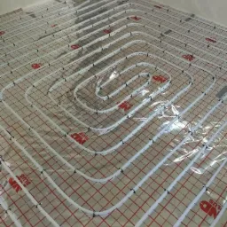 Rozłożone białe rury na czerwonej siatce, tworzące system ogrzewania podłogowego przed zalaniem wylewką.