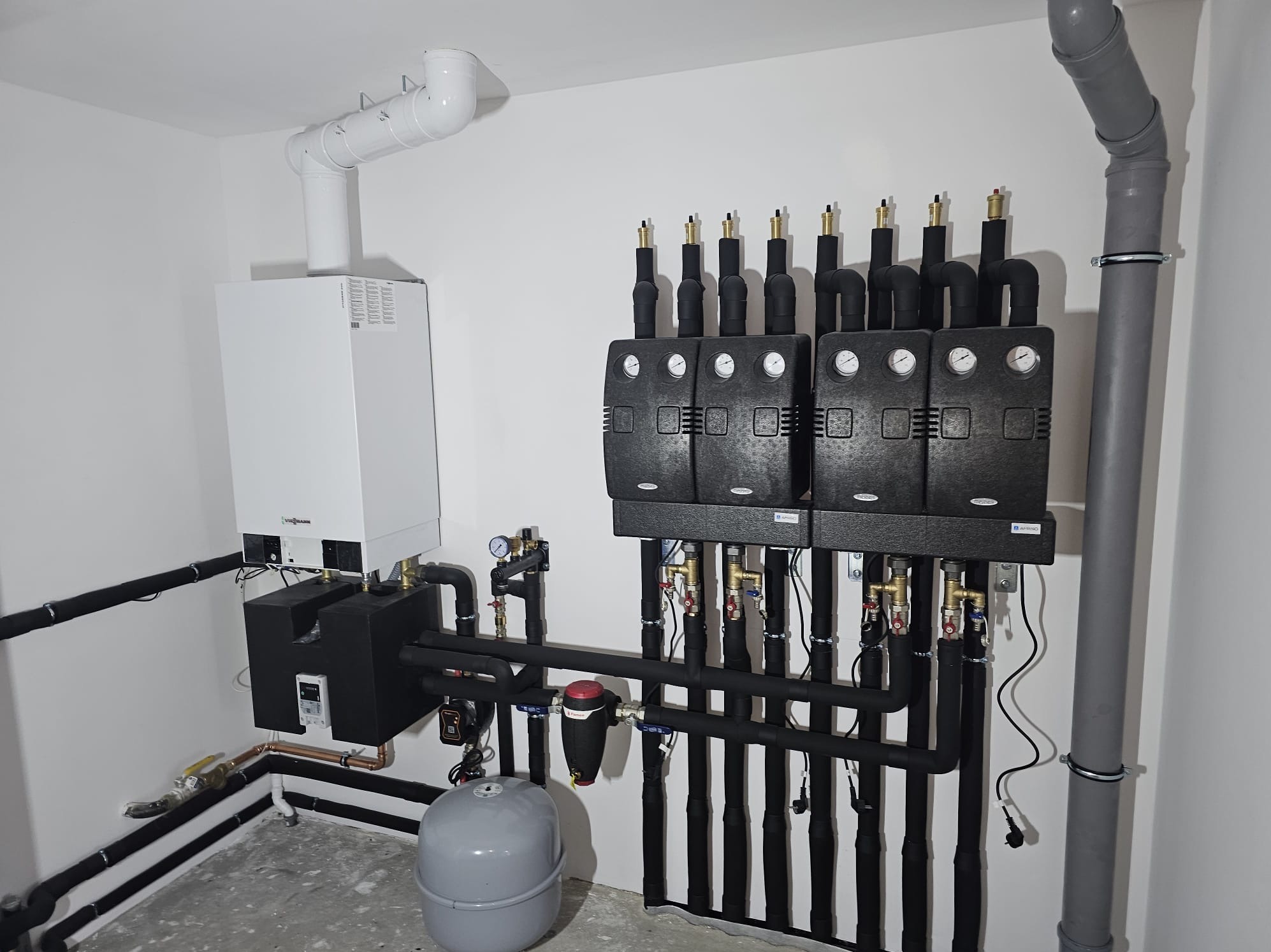 Nowoczesna kotłownia gazowa z białym piecem Viessmann, czarnymi rurami i czterema czarnymi modułami sterującymi na białej ścianie. Szara podłoga i rura odpływowa.