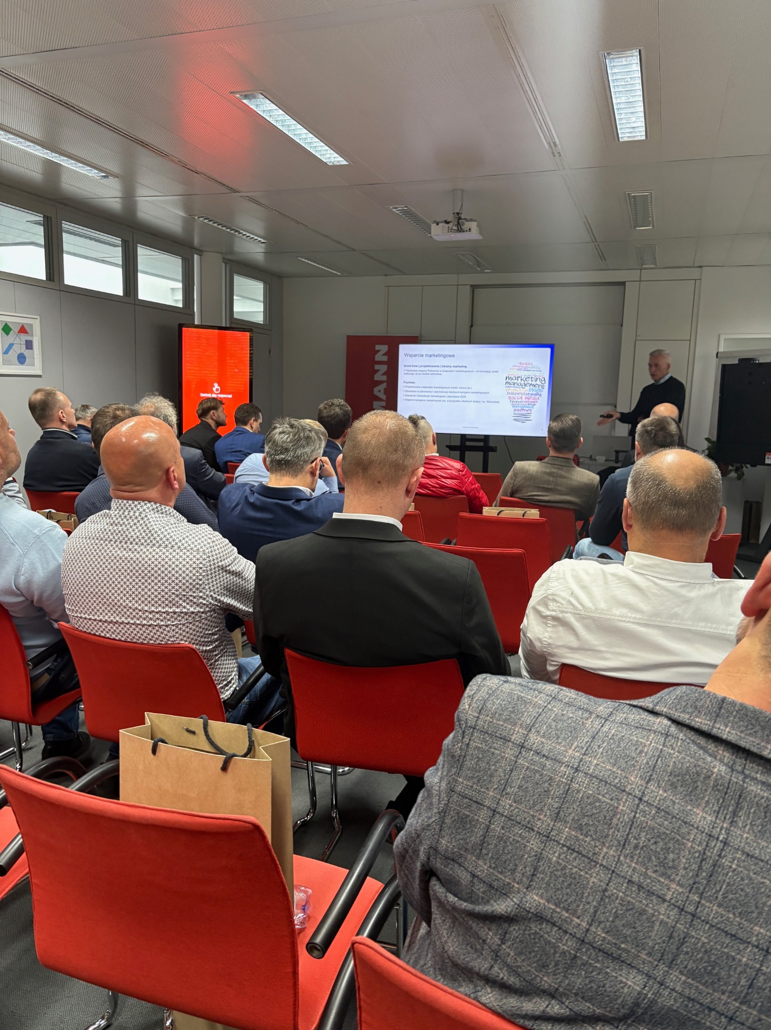 Szkolenie Viessmann: grupa instalatorów siedzi na czerwonych krzesłach, słuchając prezentacji o marketingu na ekranie. Widoczny prelegent i logo Viessmann.