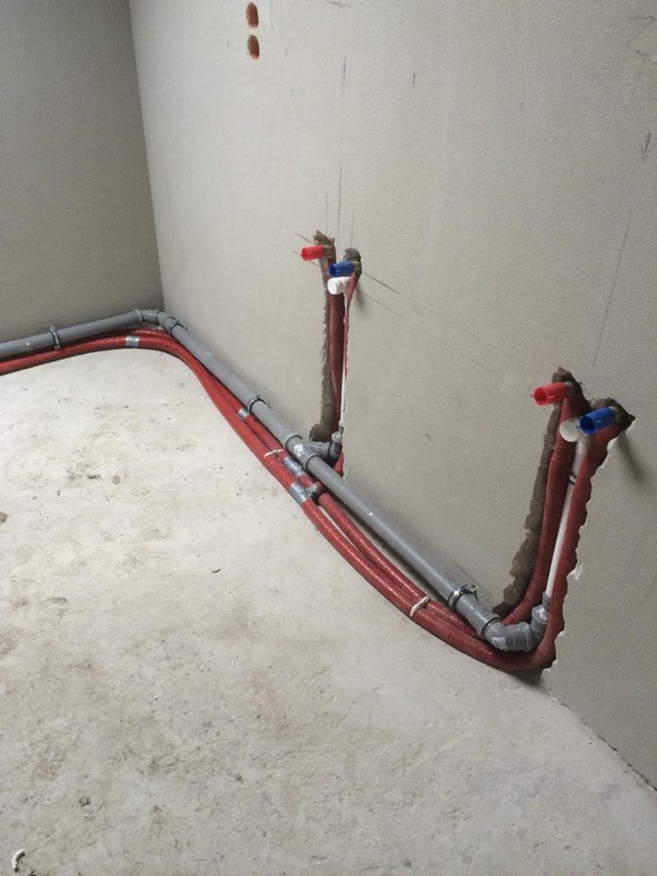 Szare i czerwone rury instalacyjne, częściowo ukryte w ścianie, z widocznymi przyłączami z czerwonymi i niebieskimi zaworami, w surowym wnętrzu z betonową podłogą i otynkowanymi ścianami.