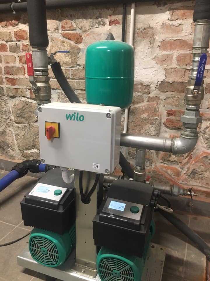 Zmontowany zestaw hydroforowy z dwoma pompami Wilo, zielonym zbiornikiem przeponowym i szarą skrzynką sterowniczą na tle ściany z cegły.