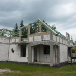 Surowy stan domu z białego bloczka, z widoczną konstrukcją dachu i żurawiem budowlanym w tle. Elementy architektoniczne w stylu łukowym. Etap budowy.