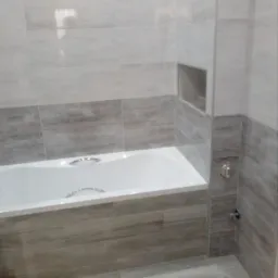 Wykończona łazienka z wanną obudowaną płytkami imitującymi drewno, szare płytki na ścianach i podłodze, oraz wnękami w ścianie.