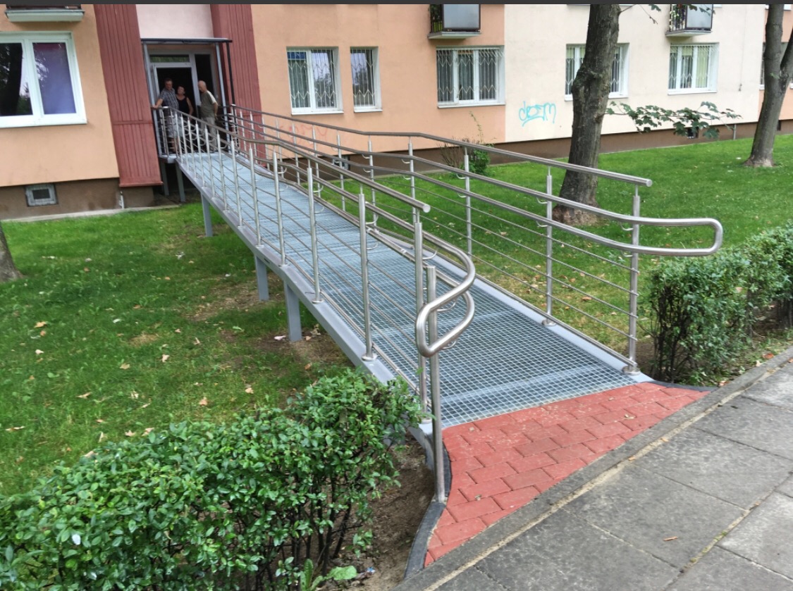 Zewnętrzny podjazd dla osób z niepełnosprawnościami, z metalową kratownicą i stalowymi poręczami, prowadzący do wejścia budynku wielorodzinnego, otoczony zielenią.