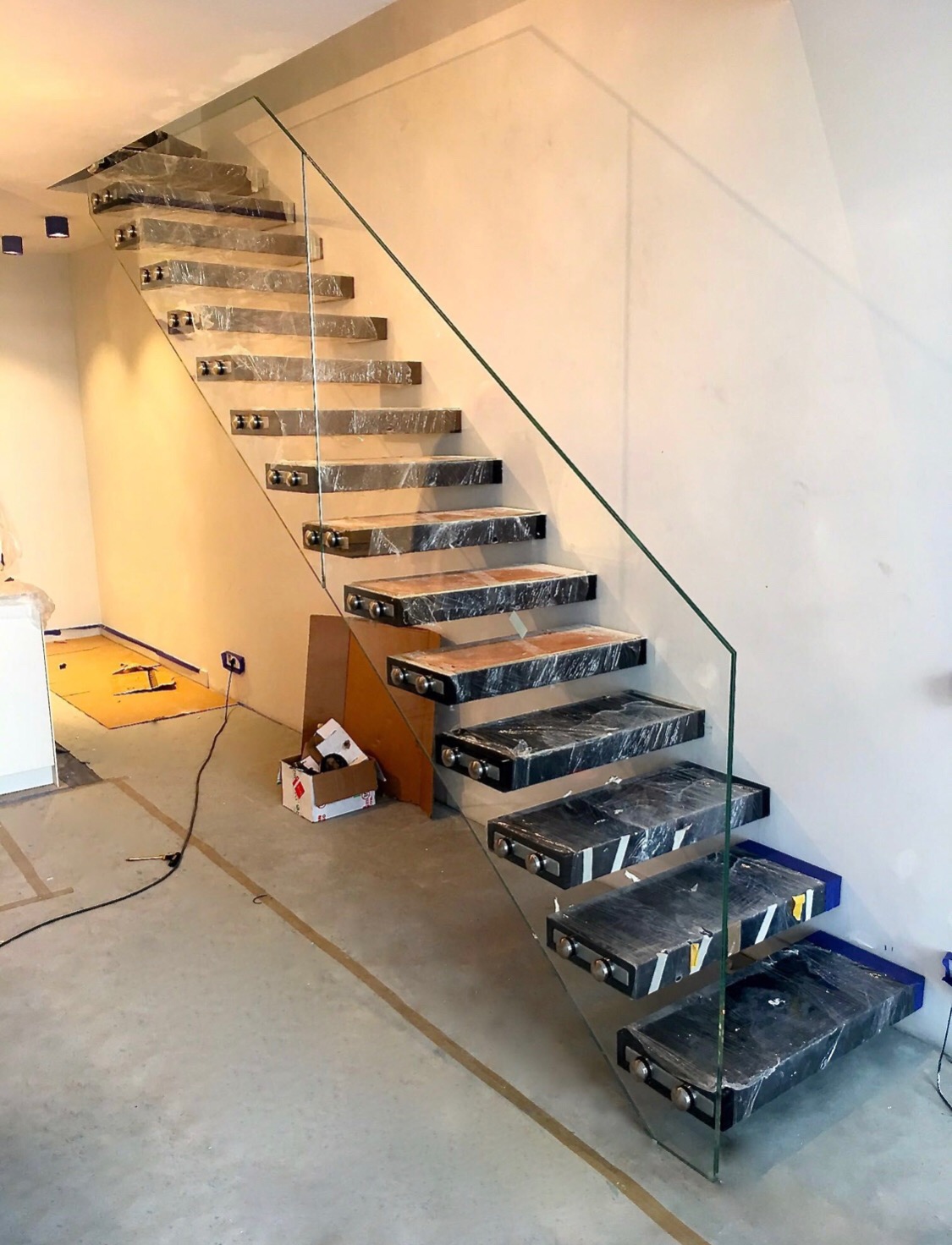 Szklana balustrada z marmurowymi schodami w trakcie montażu, zabezpieczone folią, widoczne narzędzia i karton na podłodze.