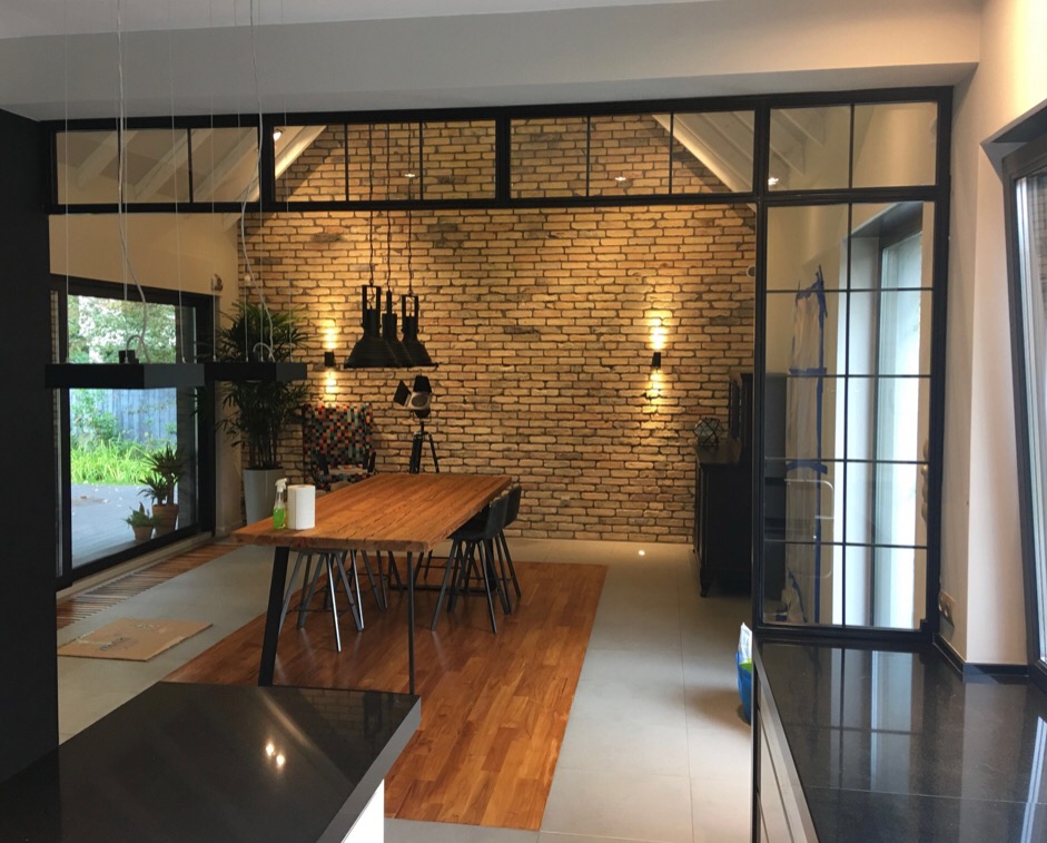 Nowoczesne wnętrze jadalni z ceglaną ścianą, drewnianym stołem na metalowych nogach, czarnymi krzesłami i przeszkloną ścianą z czarnymi szprosami.