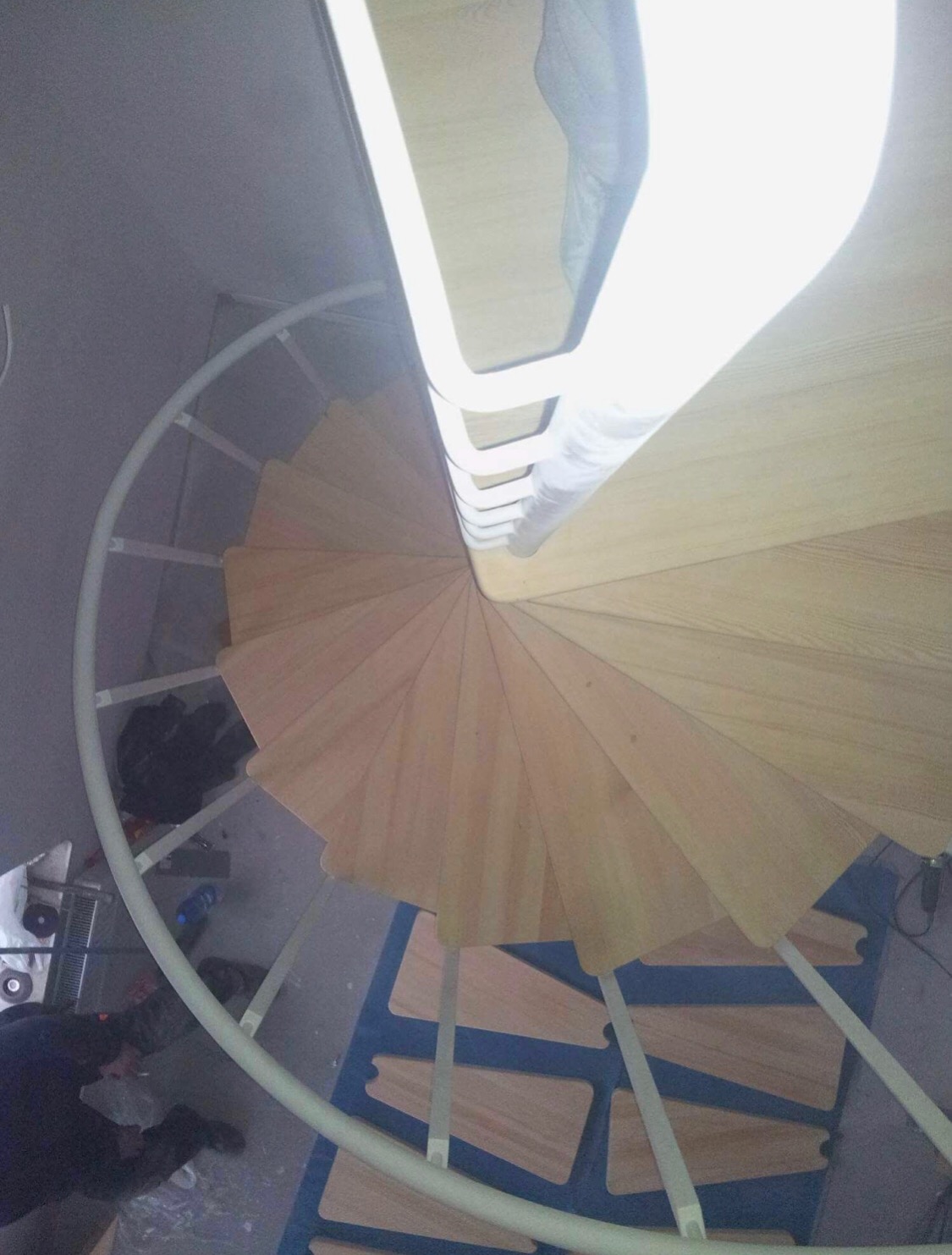 Widok z góry na spiralne schody z drewnianymi stopniami i białą poręczą, w tle widoczne narzędzia i elementy montażowe na podłodze.