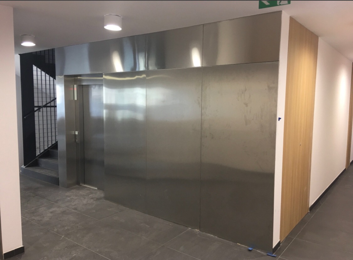 Wnętrze budynku z windą o metalowych drzwiach, widoczne schody z metalową balustradą, oświetlenie sufitowe i panele ścienne z jasnego drewna.