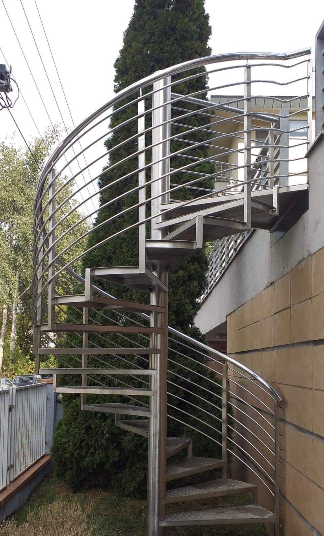 Zewnętrzne schody spiralne z balustradą ze stali nierdzewnej, widok z dołu na tle zieleni i budynku z cegły.