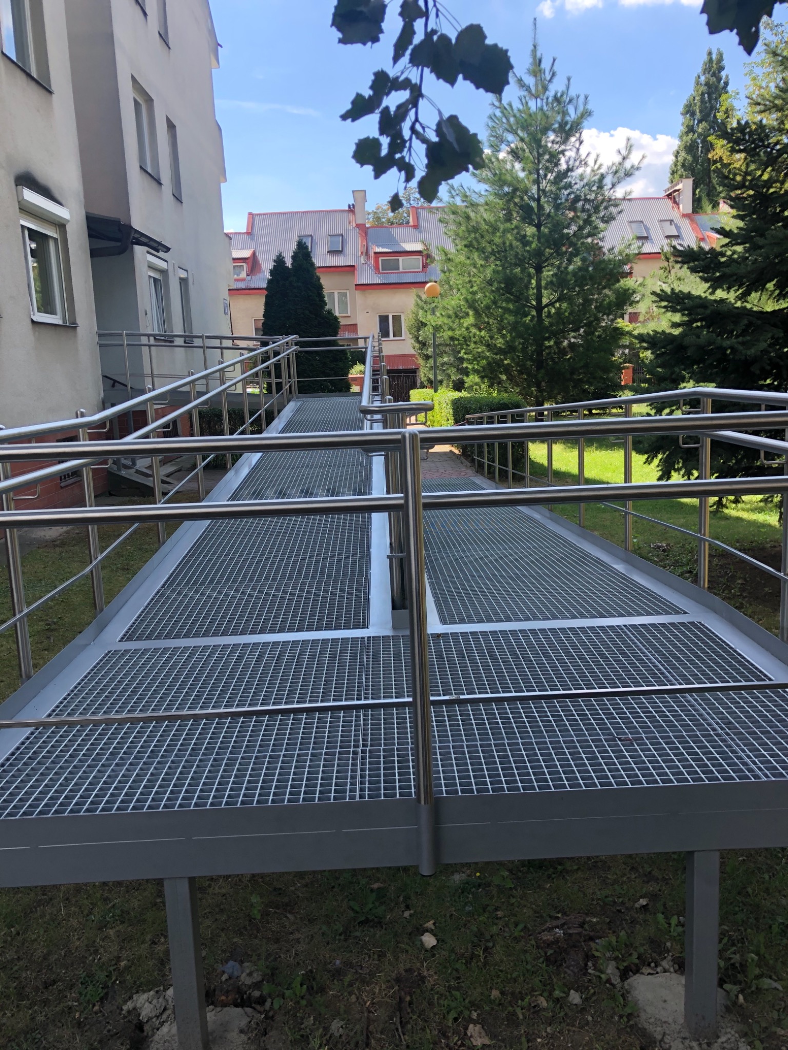 Zewnętrzna rampa dla wózków inwalidzkich z metalową kratownicą i nierdzewnymi balustradami, prowadząca do budynku mieszkalnego z widocznym fragmentem trawnika i drzew