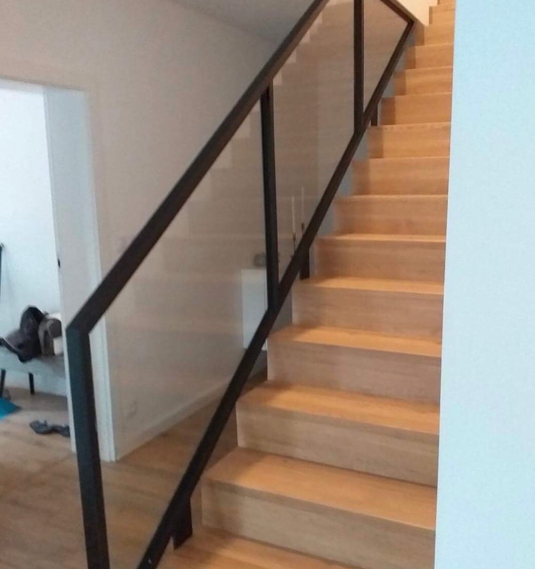 Schody drewniane z czarną, minimalistyczną poręczą i szklaną balustradą, widok z dołu schodów, jasne wnętrze.