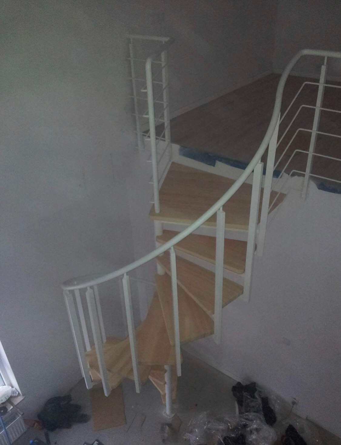 Klatka schodowa spiralna z drewnianymi stopniami i białą metalową balustradą, widok z góry, widoczne zabezpieczenia na podłodze.