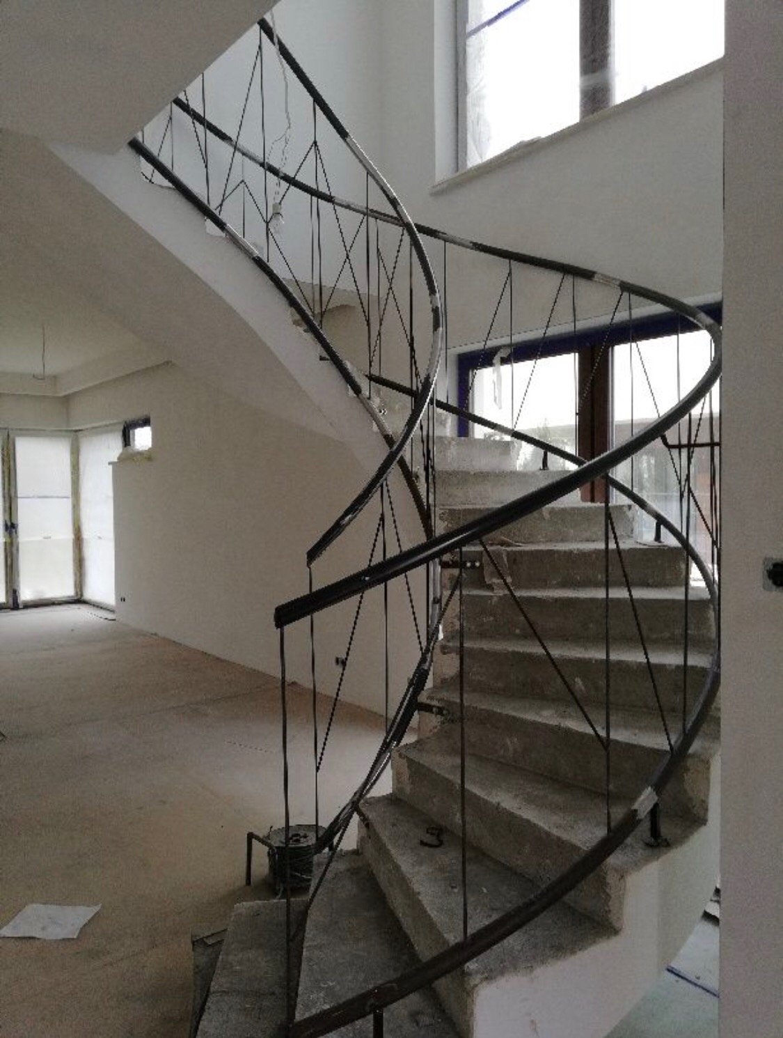 Wewnętrzna klatka schodowa w trakcie budowy z betonowymi schodami i czarną, metalową balustradą spiralną z geometrycznym wzorem wypełnienia, widok z dołu na jasne wnętrze.