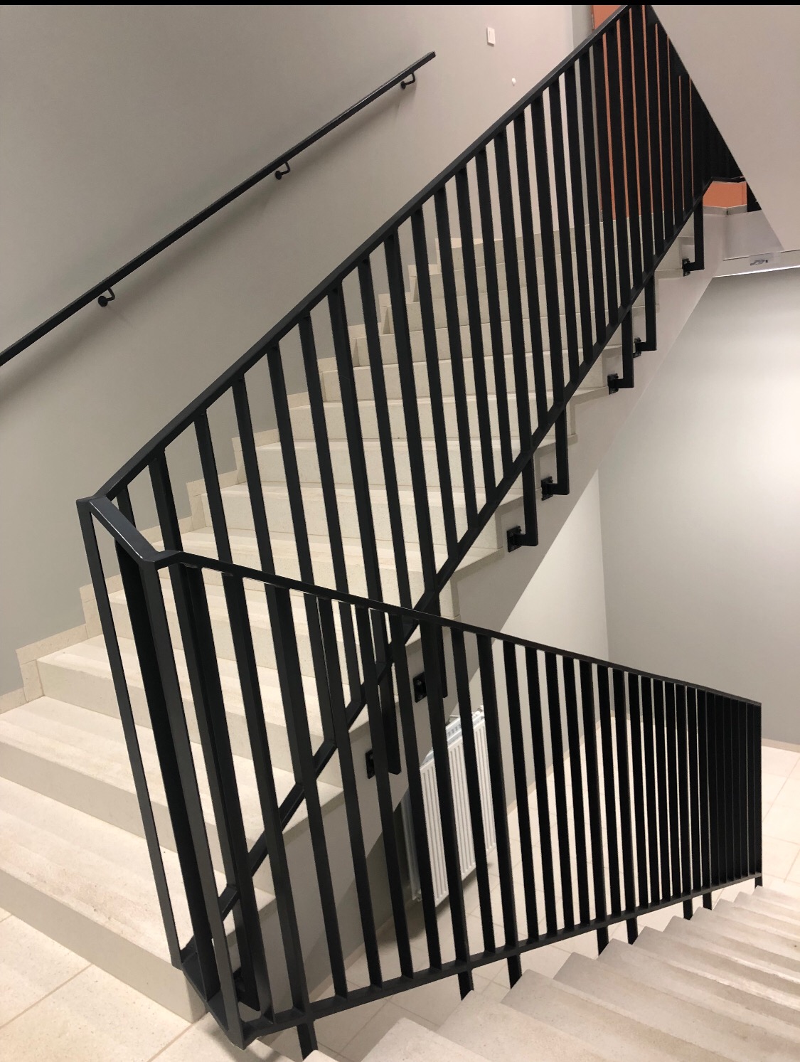 Klatka schodowa z czarną, metalową balustradą o pionowych szczeblach i poręczą zamocowaną do ściany. Białe stopnie schodów wykończone płytkami. Widoczny grzejnik za balustradą.