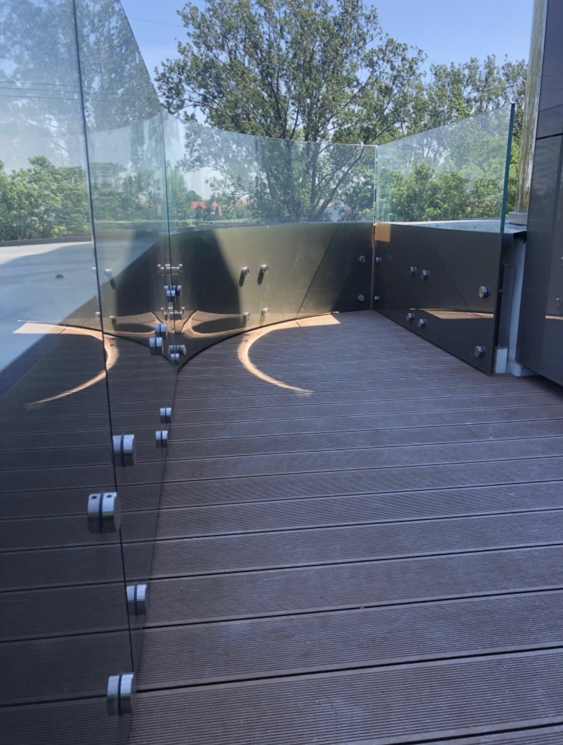 Szklana balustrada z metalowymi mocowaniami na tarasie z desek kompozytowych, widok na zieleń w tle.