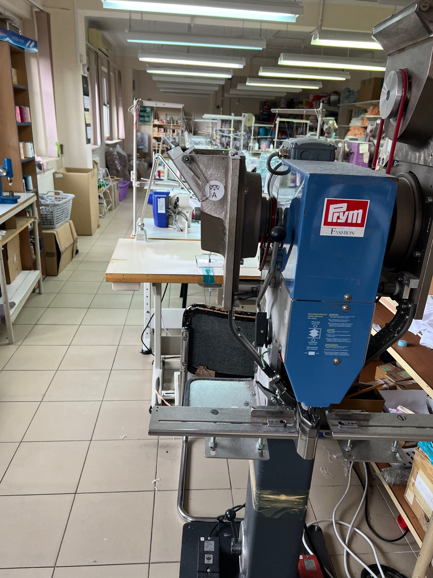 Wnętrze szwalni z widoczną maszyną do szycia z logo FRYM Fashion, w tle inne maszyny i stanowiska pracy, oświetlenie jarzeniowe.